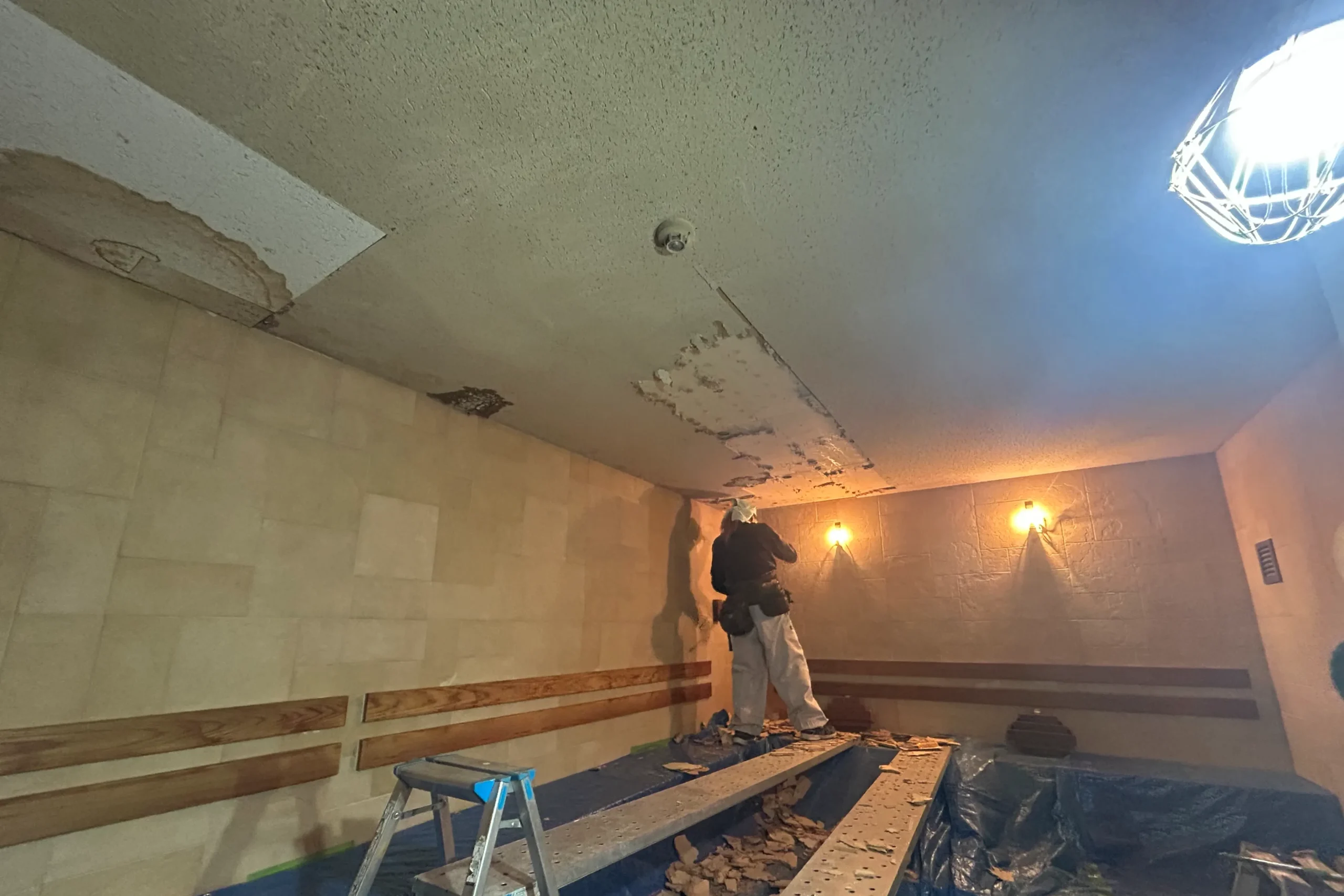 ブルーシートで養生されたサウナ室で、天井下地の撤去・交換作業を行う職人の様子。