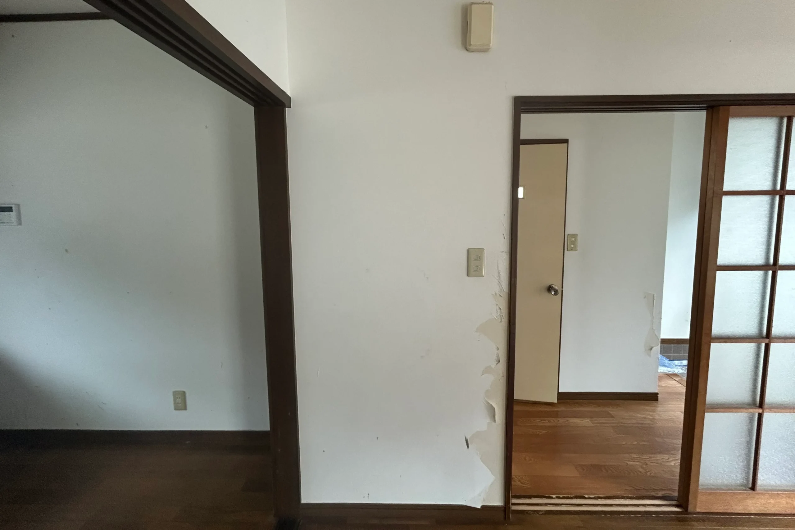 引っ越し後の空室物件にて、リビング入り口横の白い壁紙が広範囲に剥がれ、下地の石膏ボードが露出している様子。