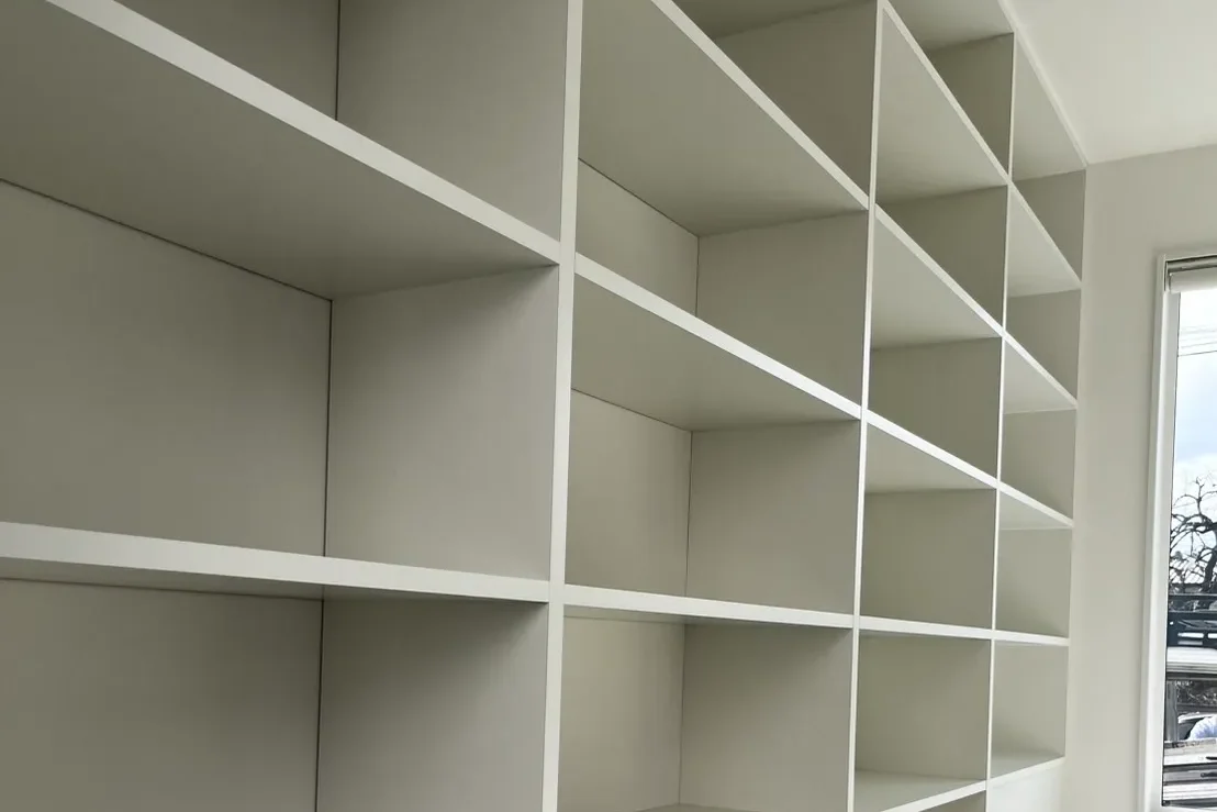 窓の左側に設置された、多段構成の白い壁面本棚。部屋の角まで隙間なくフィットしている様子。